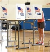 A woman standing at a voter privacy box. She's filling out a ballot in a school gym.